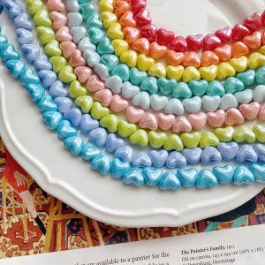 Puede incluir: Una colección de cuentas en forma de corazón en un arcoíris de colores, incluyendo azul, verde, amarillo, naranja y rojo. Las cuentas están dispuestas en filas curvas sobre un plato blanco, creando una exhibición vibrante.