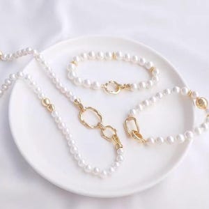 Puede incluir: Tres collares de perlas con detalles dorados. Los collares están sobre un plato blanco. Los collares tienen diferentes longitudes y estilos de detalles dorados.