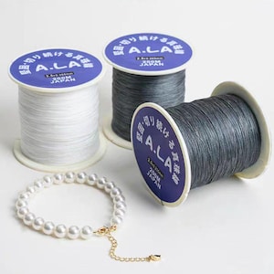 以下が含まれることがあります： 白、グレー、濃いグレーの糸巻きが3つ。それぞれの糸巻きには「A.LA」と「200M JAPAN」と書かれた日本語のラベルが付いています。