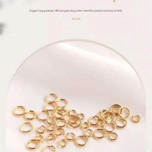 Puede incluir: Primer plano de un montón de pequeños anillos de salto dorados. Los anillos están esparcidos sobre una superficie blanca.  Paquete de anillos de cobre 14K oro real, larga duración de retención del color, no se desvanece fácilmente.