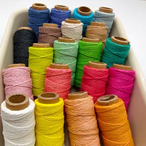 Puede incluir: Una colección de veintiuna bobinas cilíndricas de cordel artesanal multicolor dispuestas en un recipiente. Las bobinas presentan varios colores sólidos, incluyendo blanco, amarillo, naranja, melocotón, rosa, magenta, rojo, verde lima, menta, cerceta, negro, marrón, azul y gris.