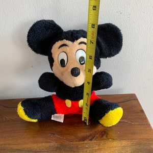 Może przedstawiać: Pluszowa zabawka Mickey Mouse w kolorze czarnym i ż&oacute;łtym siedząca na drewnianej powierzchni. Zabawka ma około 18 cm wysokości.