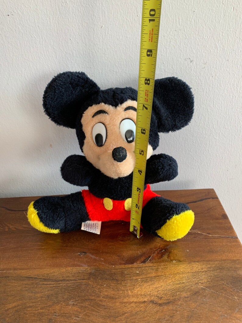 Może przedstawiać: Pluszowa zabawka Mickey Mouse w kolorze czarnym i ż&oacute;łtym siedząca na drewnianej powierzchni. Zabawka ma około 18 cm wysokości.
