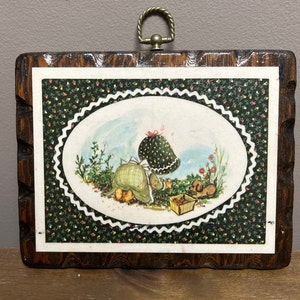 May include: A wooden plaque with a picture of a girl in a green dress and a red hat picking strawberries with a dog. The plaque has a floral pattern border and a brass hanging loop.
