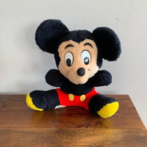 Może przedstawiać: Pluszowa zabawka Mickey Mouse w kolorze czarnym i brązowym z dużymi oczami i czerwono-ż&oacute;łtym strojem. Zabawka siedzi na drewnianej powierzchni.