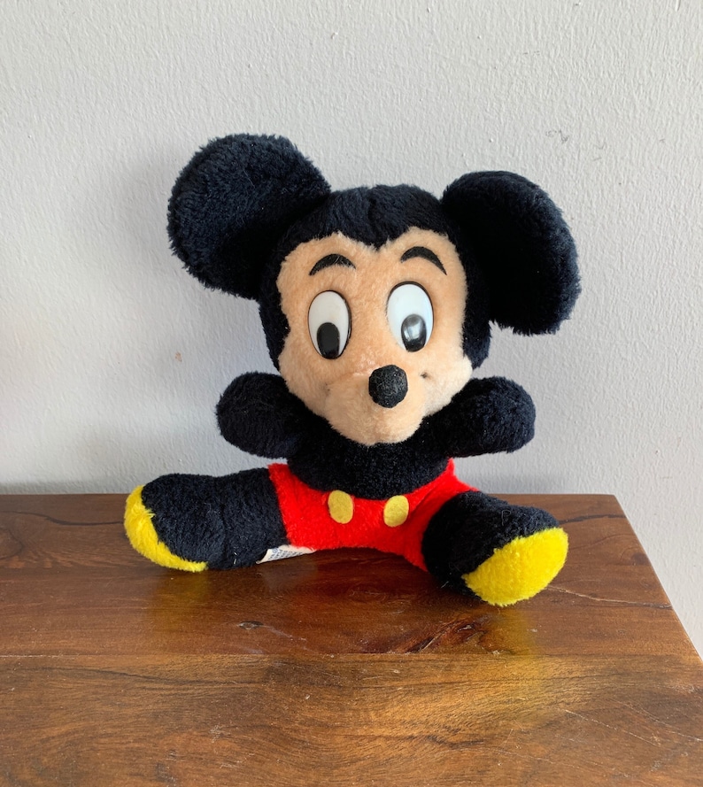 Może przedstawiać: Pluszowa zabawka Mickey Mouse w kolorze czarnym i brązowym z dużymi oczami i czerwono-ż&oacute;łtym strojem. Zabawka siedzi na drewnianej powierzchni.