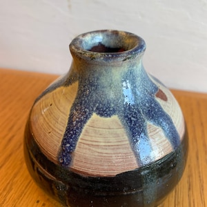 以下が含まれることがあります： 口が広く、丸い小さな陶器の花瓶。花瓶は青と白の釉薬で装飾されており、底に黒い帯が巻かれている。