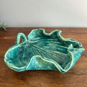 May include: A teal ceramic dish shaped like a large leaf with a glossy finish. The dish features raised, wavy edges and visible vein details embossed across the surface, mimicking the natural structure of a plant leaf with a curved stem handle.