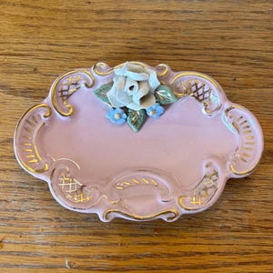 Puede incluir: Un pequeño plato de porcelana rosa con ribete dorado y un diseño floral en relieve. El plato tiene un borde festoneado y un delicado diseño floral.