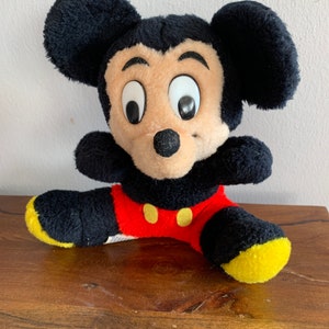Może przedstawiać: Pluszowa zabawka Mickey Mouse siedząca ze skrzyżowanymi nogami. Zabawka jest czarna, brązowa, czerwona i ż&oacute;łta. Mickey Mouse ma duże czarne oczy i czarny nos.