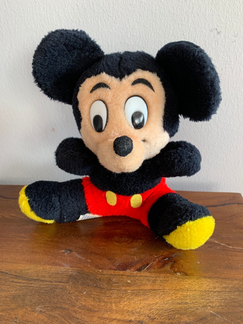 Może przedstawiać: Pluszowa zabawka Mickey Mouse siedząca ze skrzyżowanymi nogami. Zabawka jest czarna, brązowa, czerwona i ż&oacute;łta. Mickey Mouse ma duże czarne oczy i czarny nos.