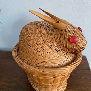 May include: A woven wicker basket shaped like a rabbit. The basket has a round body with a lid and a flared base. It features wooden ears and red glass bead eyes. The basket is light brown and sits on a wooden surface.