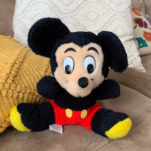 Może przedstawiać: Pluszowa zabawka Mickey Mouse w kolorze czarnym i czerwonym z ż&oacute;łtymi akcentami na uszach i stopach. Zabawka ma czarny nos i duże białe oczy.