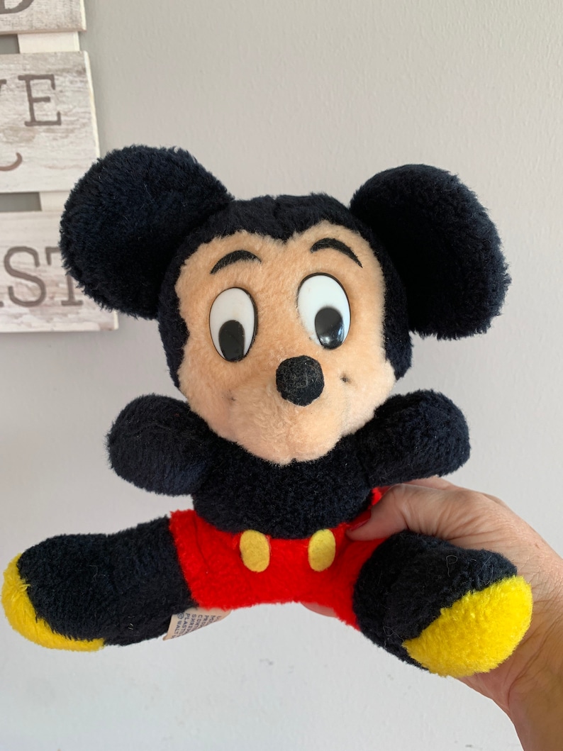 Może przedstawiać: Pluszowa zabawka vintage Mickey Mouse z czarnym futerkiem, dużymi białymi oczami i czerwono-ż&oacute;łtym strojem.