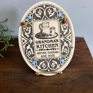 May include: An oval ceramic wall plaque with a cream-colored background and black lace-like border patterns. The center features a graphic of a pitcher, glass, and cookies with the text "GRANDMA'S KITCHEN serves cookies and milk" in black.