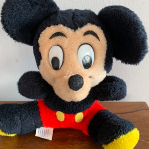 Może przedstawiać: Pluszowa zabawka Mickey Mouse w kolorze czarnym i czerwonym z dużymi oczami i ż&oacute;łtym nosem. Zabawka siedzi na drewnianej powierzchni.