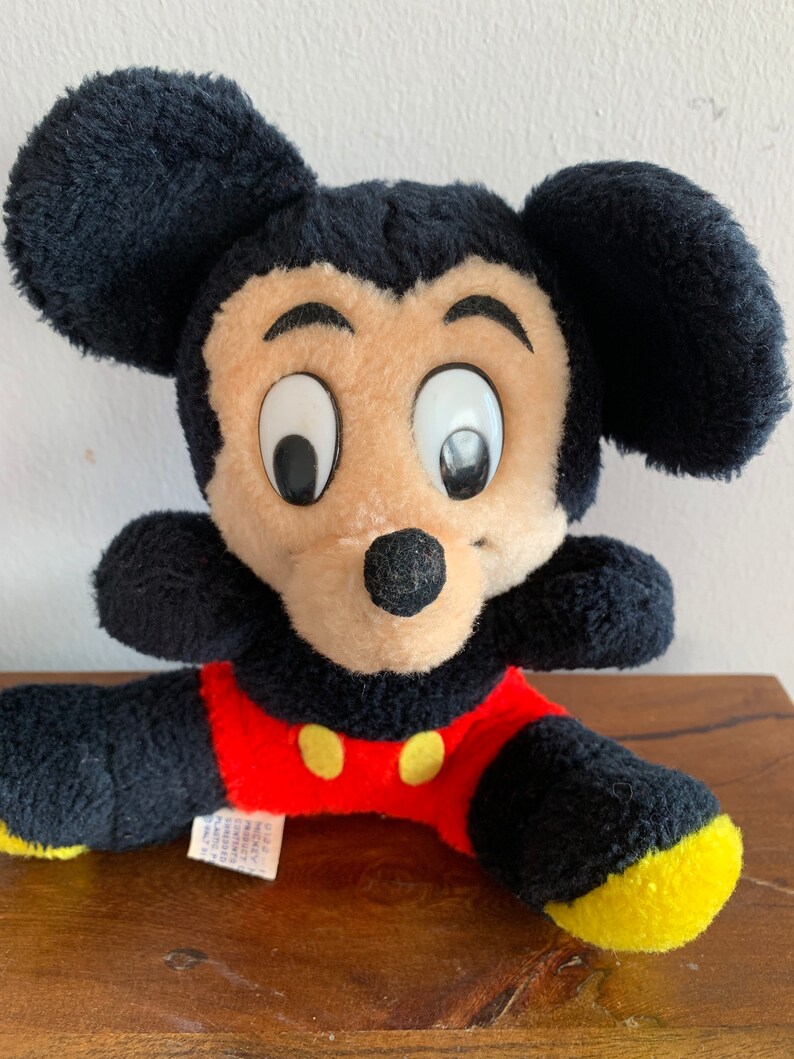 Może przedstawiać: Pluszowa zabawka Mickey Mouse w kolorze czarnym i czerwonym z dużymi oczami i ż&oacute;łtym nosem. Zabawka siedzi na drewnianej powierzchni.