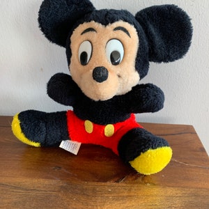 Może przedstawiać: Pluszowa zabawka Mickey Mouse w kolorze czarnym i czerwonym z ż&oacute;łtymi butami. Zabawka siedzi na drewnianej powierzchni.