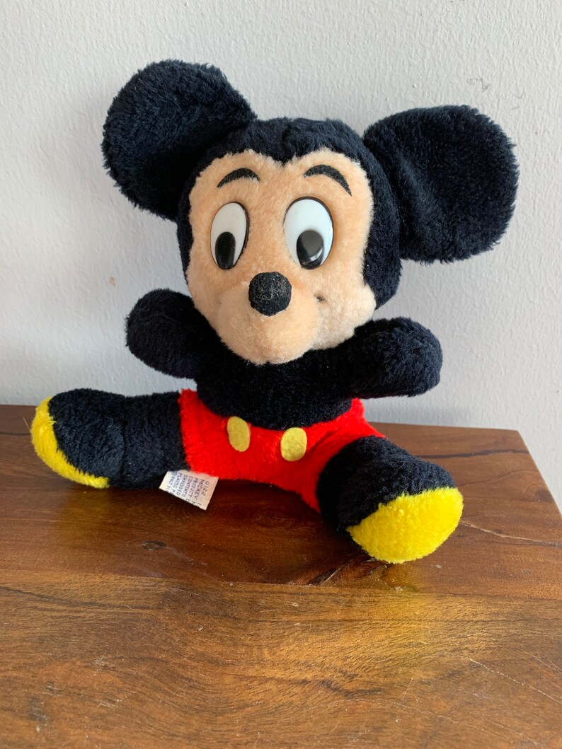 Może przedstawiać: Pluszowa zabawka Mickey Mouse w kolorze czarnym i czerwonym z ż&oacute;łtymi butami. Zabawka siedzi na drewnianej powierzchni.