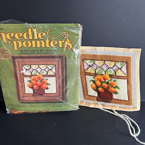 May include: A needlepoint kit and finished needlepoint featuring a window scene with orange marigolds in a brown pot. The kit packaging is green with the words "Needle Pointers" and "Marigolds in the Window" visible.