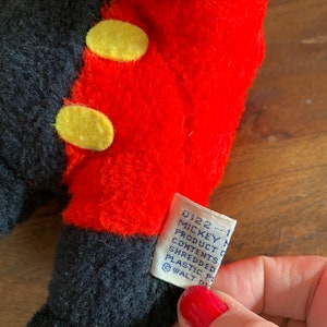 Może przedstawiać: Pluszowa zabawka Mickey Mouse w kolorze czarnym i czerwonym z ż&oacute;łtymi guzikami. Etykieta zawiera napis "0122 - 1 Mickey Mouse Product Contents Shredded Plastic Pellets &copy; WALT DISNEY"