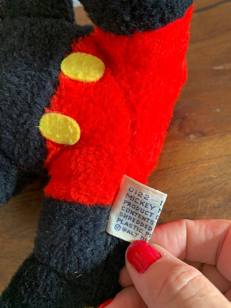 Może przedstawiać: Pluszowa zabawka Mickey Mouse w kolorze czarnym i czerwonym z ż&oacute;łtymi guzikami. Etykieta zawiera napis "0122 - 1 Mickey Mouse Product Contents Shredded Plastic Pellets &copy; WALT DISNEY"