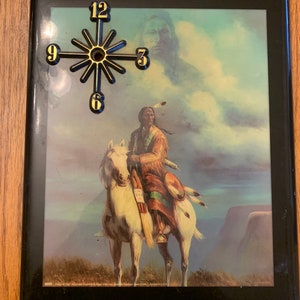 May include: A framed wall clock with a black frame and a black and gold clock face. The clock face has a black starburst design with gold numbers and a gold hand. The image in the frame is a painting of a Native American man riding a white horse. The painting is set against a background of clouds and mountains.