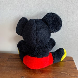Może przedstawiać: Pluszowa zabawka Mickey Mouse w kolorze czarnym siedząca na drewnianej powierzchni. Zabawka ma na sobie czerwone spodenki z ż&oacute;łtymi akcentami.