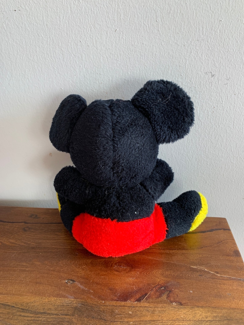 Może przedstawiać: Pluszowa zabawka Mickey Mouse w kolorze czarnym siedząca na drewnianej powierzchni. Zabawka ma na sobie czerwone spodenki z ż&oacute;łtymi akcentami.
