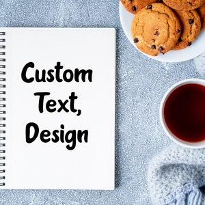 Peut inclure: Un carnet blanc avec une reliure spirale noire et le texte "Custom Text, Design" écrit en noir sur la couverture. Le carnet est posé sur une surface texturée grise avec une tasse de thé et une assiette de biscuits aux pépites de chocolat.