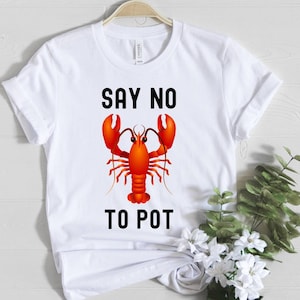 May include: White t-shirt with a bright red lobster illustration and the words "SAY NO TO POT" in black. The tee is displayed on a hanger, with the design prominently displayed on the front.