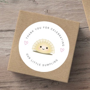 May include: White round sticker with the text "Thank you for celebrating our little dumpling" in black. A cartoon dumpling with a smiling face is in the center of the sticker. Two pink hearts are on either side of the dumpling.