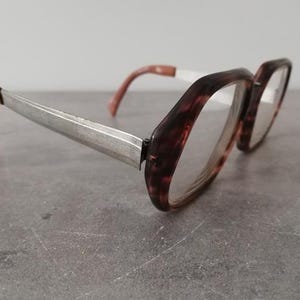 Könnte beinhalten: Eine Vintage-Brille mit Schildpattmuster auf den Rahmen. Die Bügel sind aus silberfarbenem Metall mit braunen Spitzen. Die Brille liegt auf einer grauen Oberfläche.