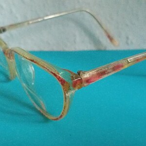 Könnte beinhalten: Eine Brille mit klaren Gläsern und einem hellfarbigen Rahmen. Der Rahmen hat ein durchscheinendes Aussehen mit roten und gelben Akzenten. Die Bügel sind dünn und silberfarben. Die Brille steht vor einem blauen Hintergrund.