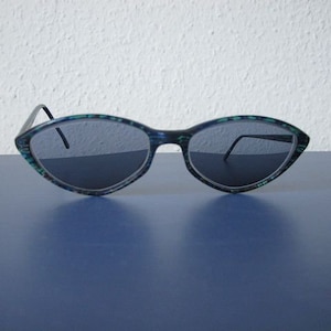 Może przedstawiać: Okulary przeciwsłoneczne typu „kocie oko” z zielono-niebieską wzorzystą oprawką i ciemnoszarymi szkłami.