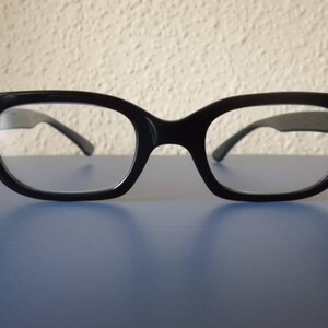 Peut inclure: Lunettes à monture noire de forme œil de chat rétro. Les lunettes ont des verres transparents et sont posées sur une surface bleue avec un arrière-plan texturé. Les montures ont de petits détails décoratifs sur les coins extérieurs.