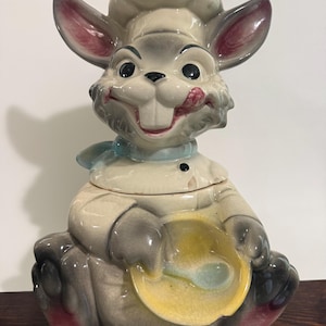 May include: A ceramic cookie jar shaped like a rabbit wearing a chef hat and uniform. The rabbit has gray fur, pink-tinted ears, and a blue neckerchief. It holds a yellow mixing bowl with a blue spoon, featuring a glossy finish and visible crazing.