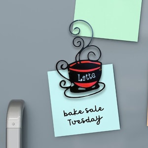May include: A light blue sticky note with the text "bake sale Tuesday" on a gray refrigerator. A black metal coffee cup magnet with the word "Latte" written on it is attached to the note.