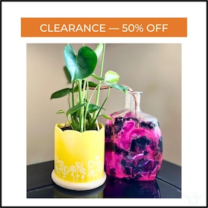 May include: A yellow ceramic planter with a scalloped rim and white floral design, holding a green plant. Next to it is a clear glass bottle with pink and black swirls. Text at the top reads "CLEARANCE - 50% OFF".