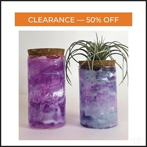 May include: Two cylindrical glass jars with cork stoppers. The jars have a marbled purple and white design. One jar contains a plant. The text "CLEARANCE - 50% OFF" is in an orange banner at the top of the image.