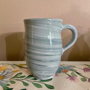 May include: A ceramic mug featuring a marbled pattern of white and light gray horizontal swirls. The mug has a cylindrical body with a slightly tapered base and a curved handle on the right side. The surface has a smooth, glossy finish throughout.