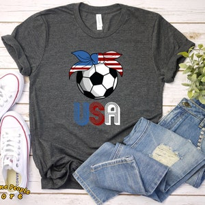Puede incluir: Una camiseta gris oscura con un balón de fútbol que lleva una bandana roja, blanca y azul. La palabra "USA" está impresa debajo del balón de fútbol en una fuente de estilo retro.