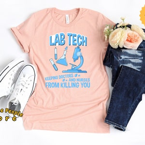 May include: A pink t-shirt with the text "Lab Tech Keeping Doctors and Nurses From Killing You" in blue and black text. The shirt also features a blue beaker and microscope graphic.
