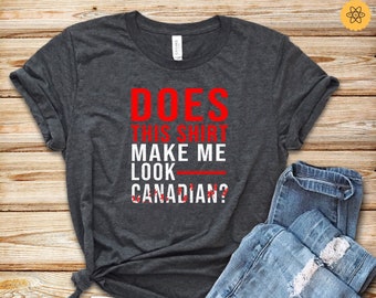 May include: A dark grey t-shirt with the text "DOES THIS SHIRT MAKE ME LOOK CANADIAN?" in red. The shirt is laid flat on a wooden surface with a pair of blue jeans and white trainers.