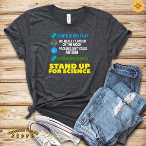 May include: A dark grey t-shirt with the text "EARTH IS NOT FLAT", "WE REALLY LANDED ON THE MOON", "VACCINES DON'T CAUSE AUTISM", "EVOLUTION IS FACT", and "STAND UP FOR SCIENCE" in white and yellow text.