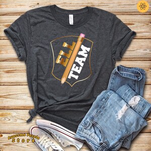 May include: A dark gray t-shirt with a gold shield design featuring a pencil and the text "ELL TEAM".