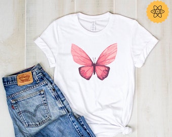 Op de afbeelding: Een wit T-shirt met een roze vlinderprint. De vlinder heeft een aquareleffect en is gecentreerd op het shirt. Het shirt ligt op een houten oppervlak met een blauwe spijkerbroek.