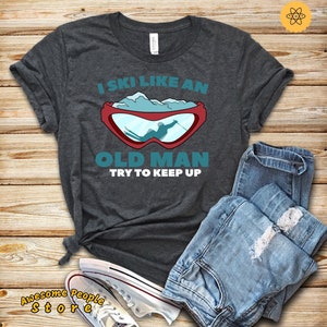 Könnte beinhalten: Ein dunkelgraues T-Shirt mit dem Text "I ski like an old man try to keep up" und einer Grafik eines Skifahrers mit einer Skibrille.
