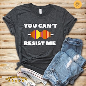 Puede incluir: Una camiseta gris oscuro con el texto "YOU CAN'T RESIST ME" en blanco. Una resistencia colorida se representa entre las palabras "YOU" y "RESIST".