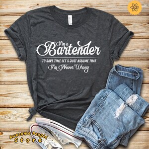 Pode incluir: Uma camiseta cinza escura com texto branco que diz "I'm a Bartender To save time let's just assume that I'm Never Wrong". A camiseta está dobrada e colocada sobre uma superfície de madeira com um par de calças jeans azuis e tênis brancos.